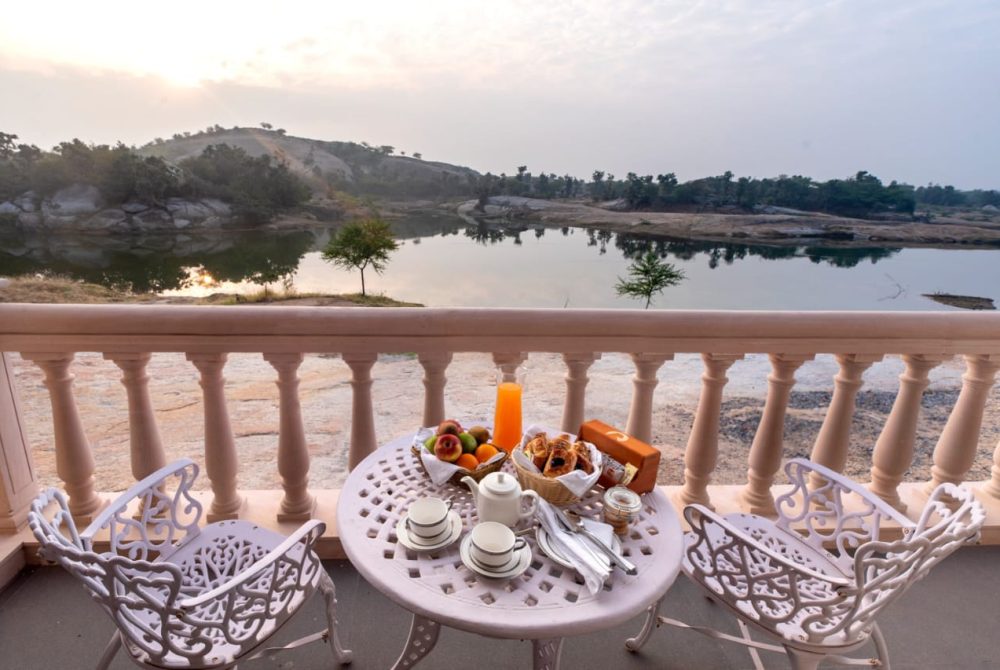Breakfast setup on a white wrought-iron balcony table beside a calm river at sunrise.
