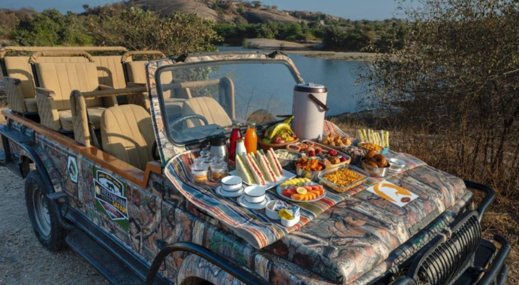 Open-top jeep with camouflage wrap parked beside a lake; the hood serves as a spread of sandwiches, fruit, pastries, drinks and a coffee pot for a roadside picnic.