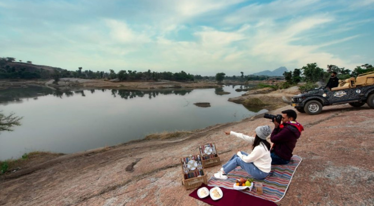 Couple picnicking on a striped blanket by a calm river, with snacks and fruit laid out; photographer takes their photo while a rugged jeep sits on the rocky shore nearby.