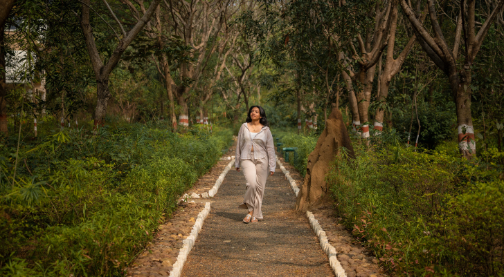 Woman in light beige outfit walking along a tree-lined park path, looking ahead as she strolls.