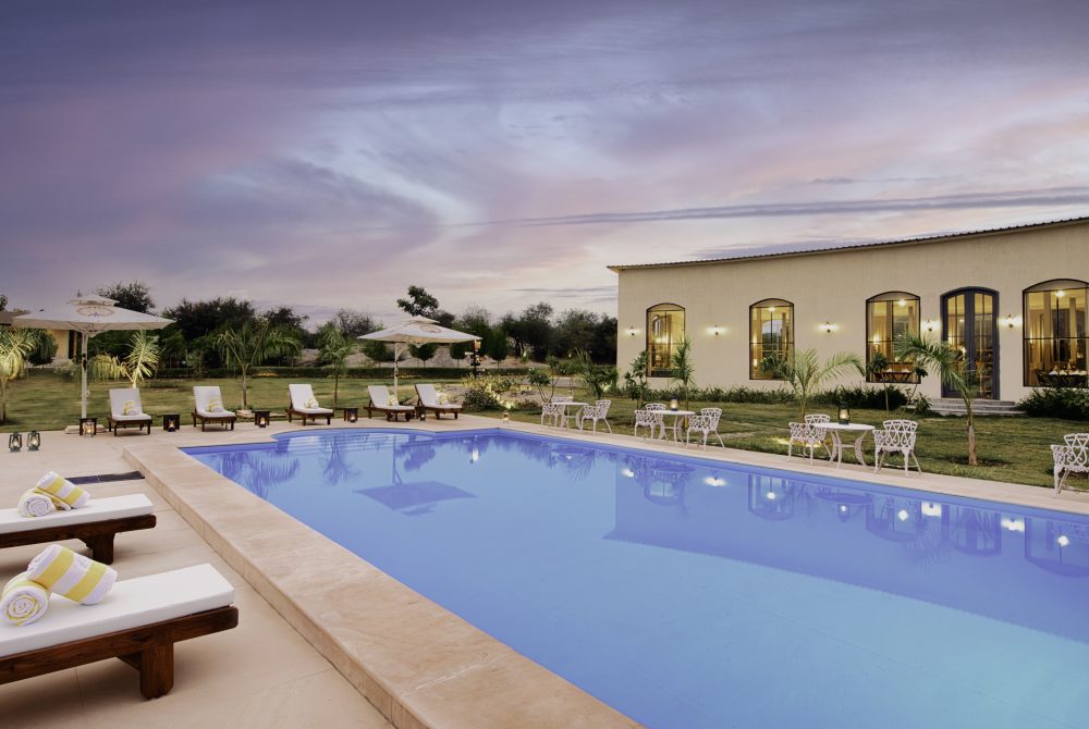 Luxury hotel pool area at dusk with white lounge chairs, umbrellas, and a beige building with arched windows