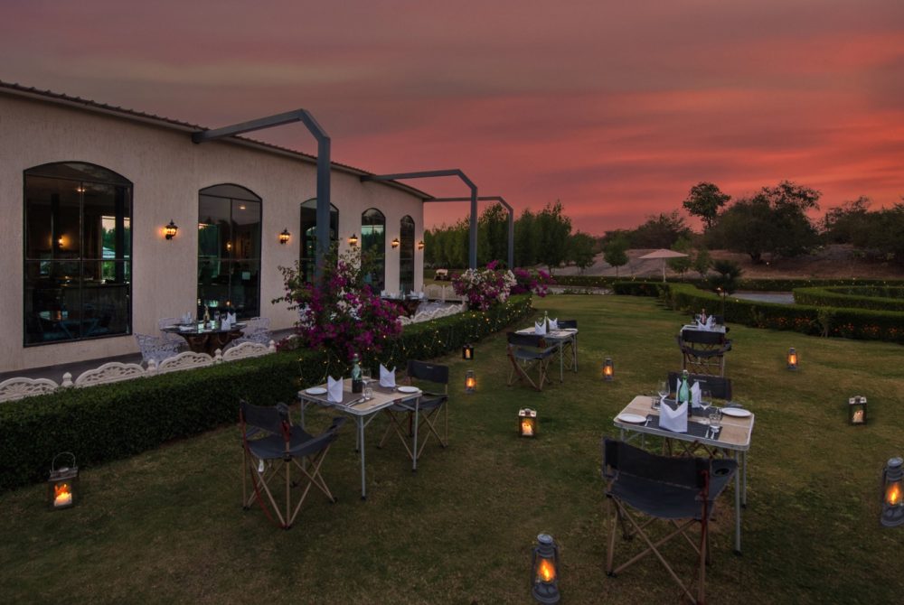 Outdoor dining setup at dusk along a beige building, with string lights, lanterns, and set tables on a lawn under a pink-orange sunset.