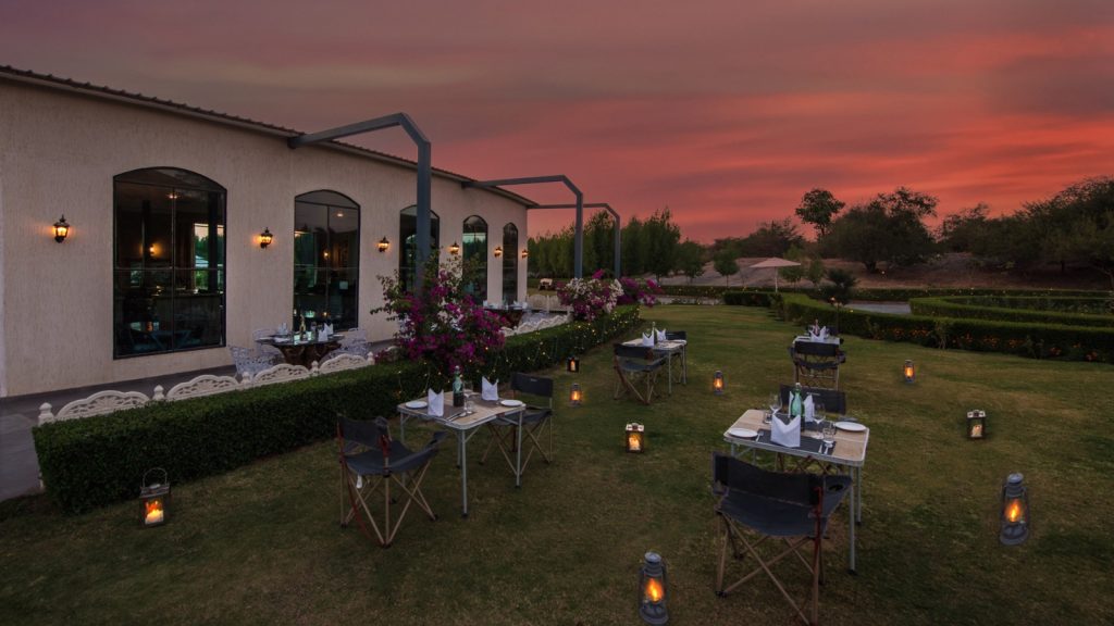 Outdoor dining setup at dusk along a beige building, with string lights, lanterns, and set tables on a lawn under a pink-orange sunset.