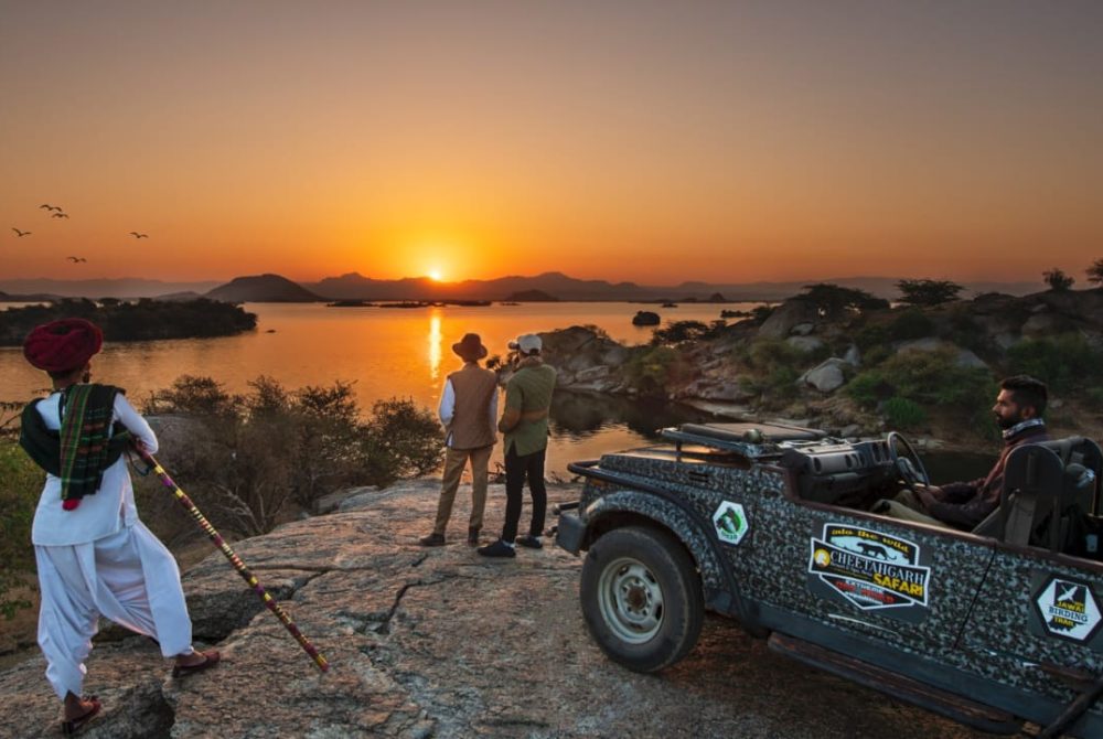 Group of three men and a driver on a rocky overlook at sunset, with a patterned safari jeep parked nearby.