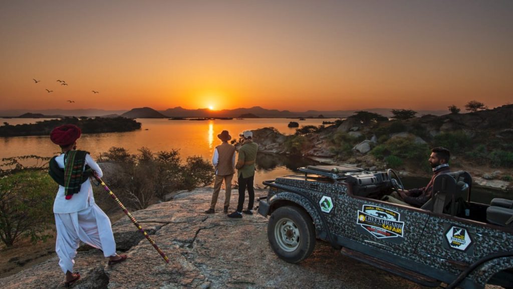 Group of three men and a driver on a rocky overlook at sunset, with a patterned safari jeep parked nearby.