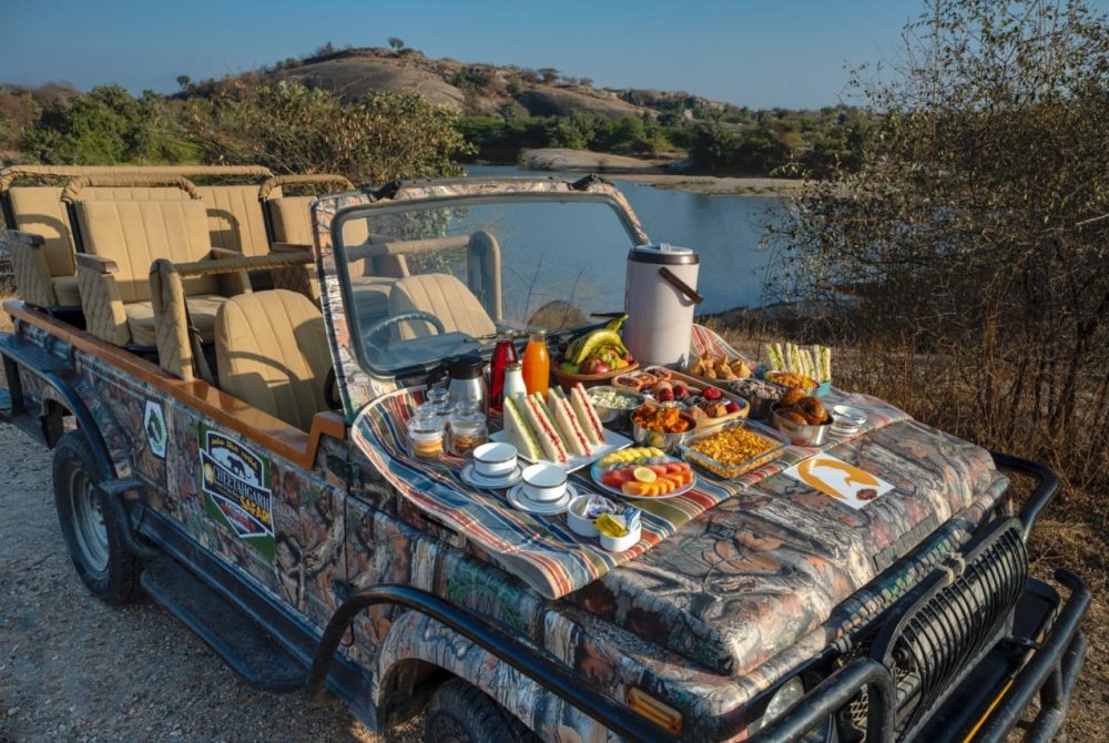 Open-top safari jeep by a lake with a table of breakfast foods laid out on the hood (sandwiches, fruit, cereals, drinks).