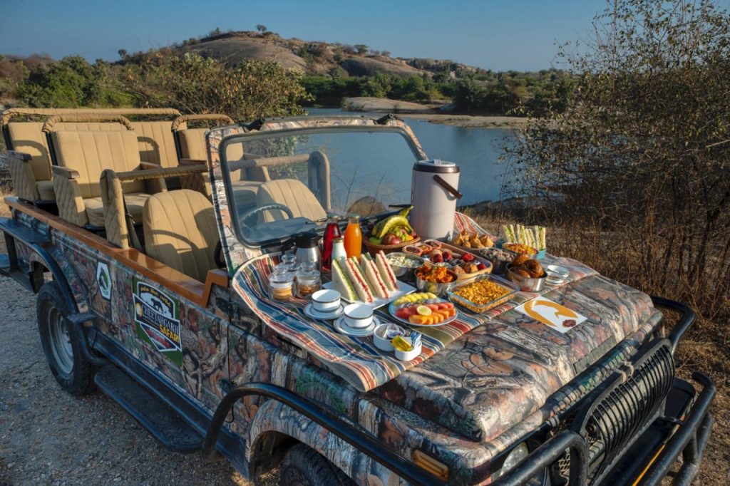Open-top safari jeep by a lake with a table of breakfast foods laid out on the hood (sandwiches, fruit, cereals, drinks).