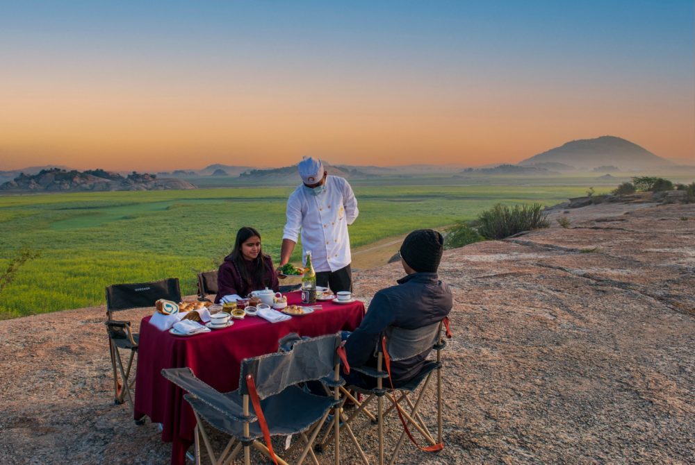 Chef in a white jacket serves food to two guests at a red-clothed table on a rocky hill, with a green valley and distant hills at sunset east sky.