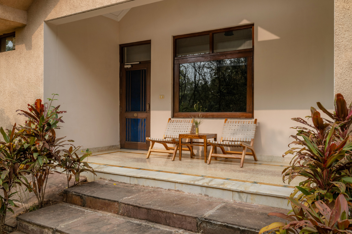Front porch of a beige house with two wooden lounge chairs and a small table between them, flanked by plants and a large window and door behind.