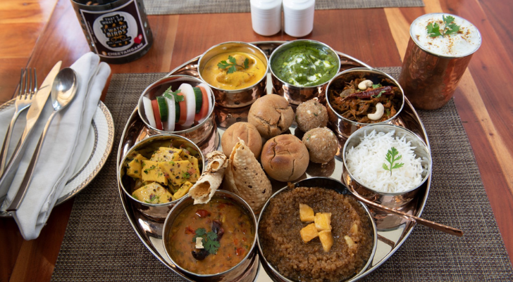 Rajasthani Theme Dinner A traditional Indian thali on a round metal tray with multiple small bowls of curries, dal, rice, breads, papad, and garnishes on a wooden table.