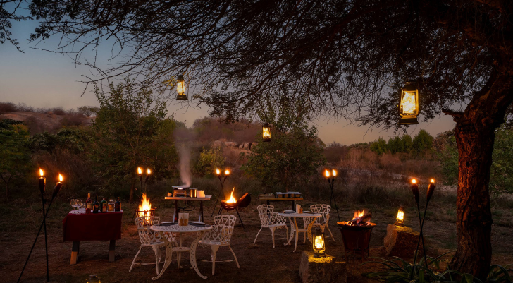 Candle Night Setup Evening outdoor dinner party with white metal chairs, tables, and torches around a fire pit on a grassy field at dusk.