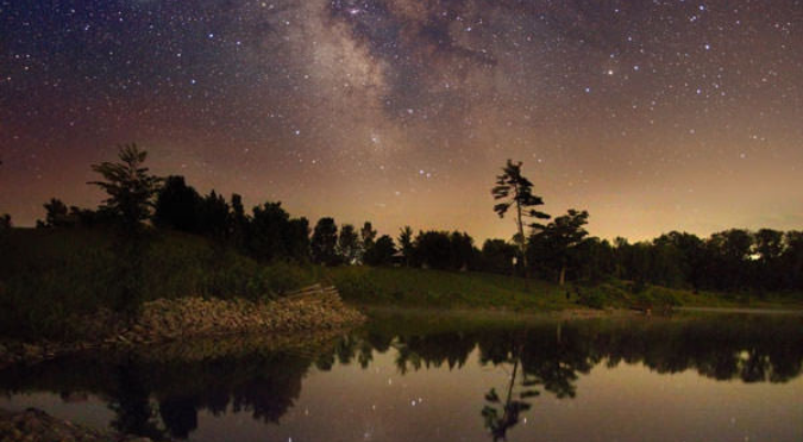 Stargazing Night sky with the Milky Way over a calm lake, silhouetted trees along the shore and a starry reflection on the water.