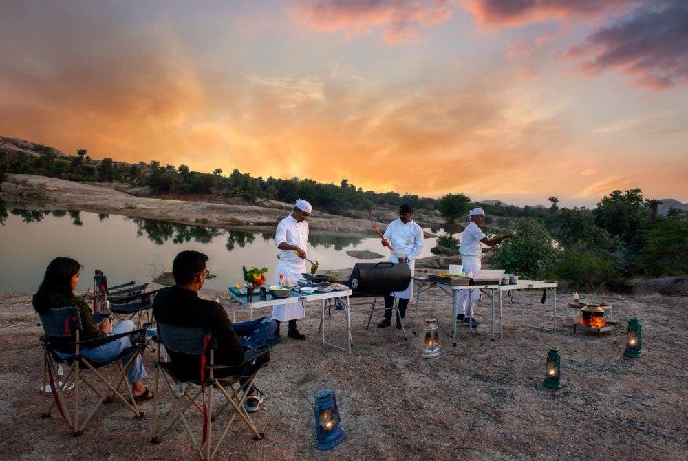 Outdoor sunset barbecue by a river: three chefs in white uniforms cook at tables with a grill while two guests sit in camping chairs and watch.