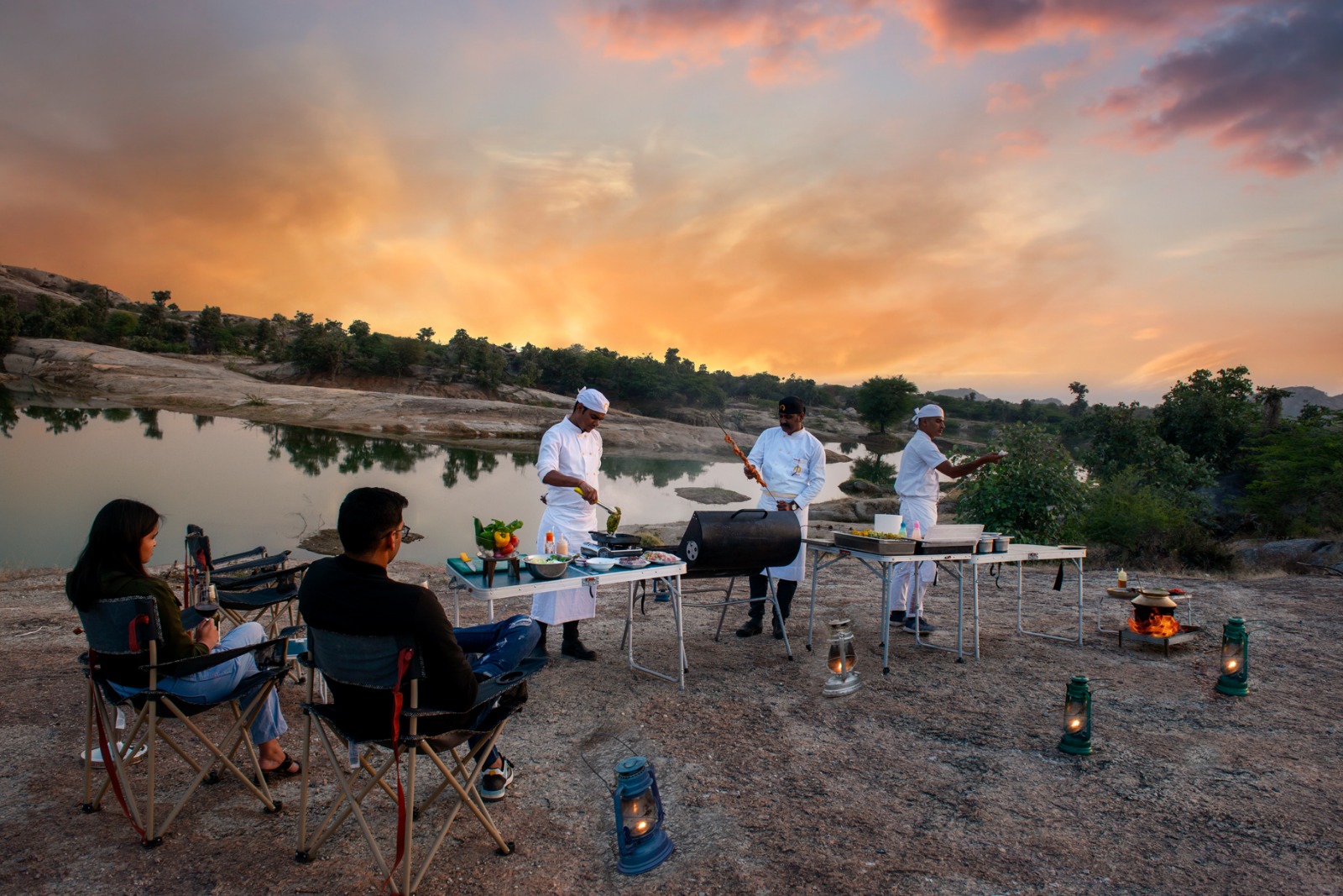 Outdoor riverside cookout at sunset: three chefs in white uniforms grill on portable stations while two diners sit in folding chairs nearby.