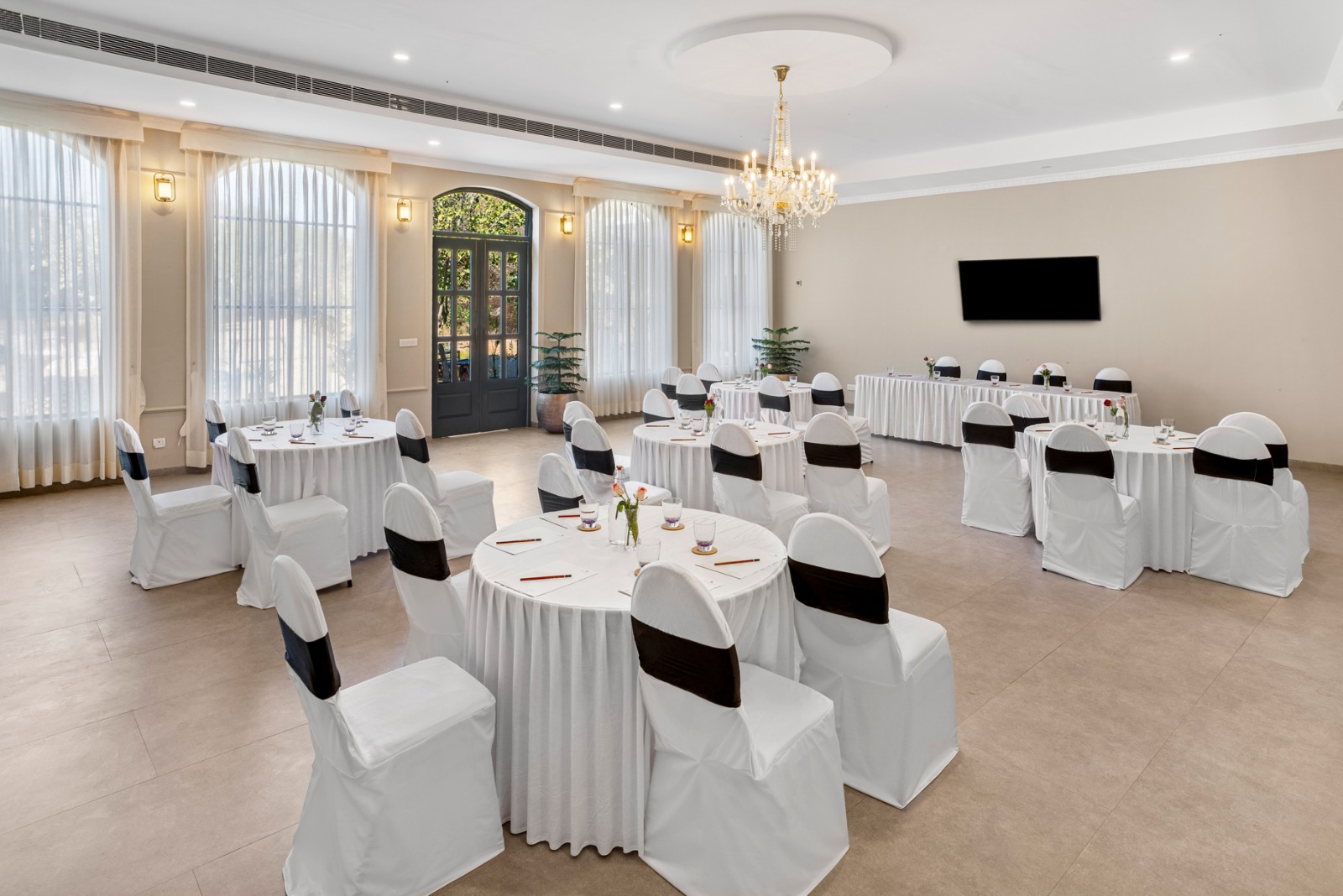 A banquet hall set for a formal event with round tables, white cloths, and chairs tied in black sashes under a chandelier.