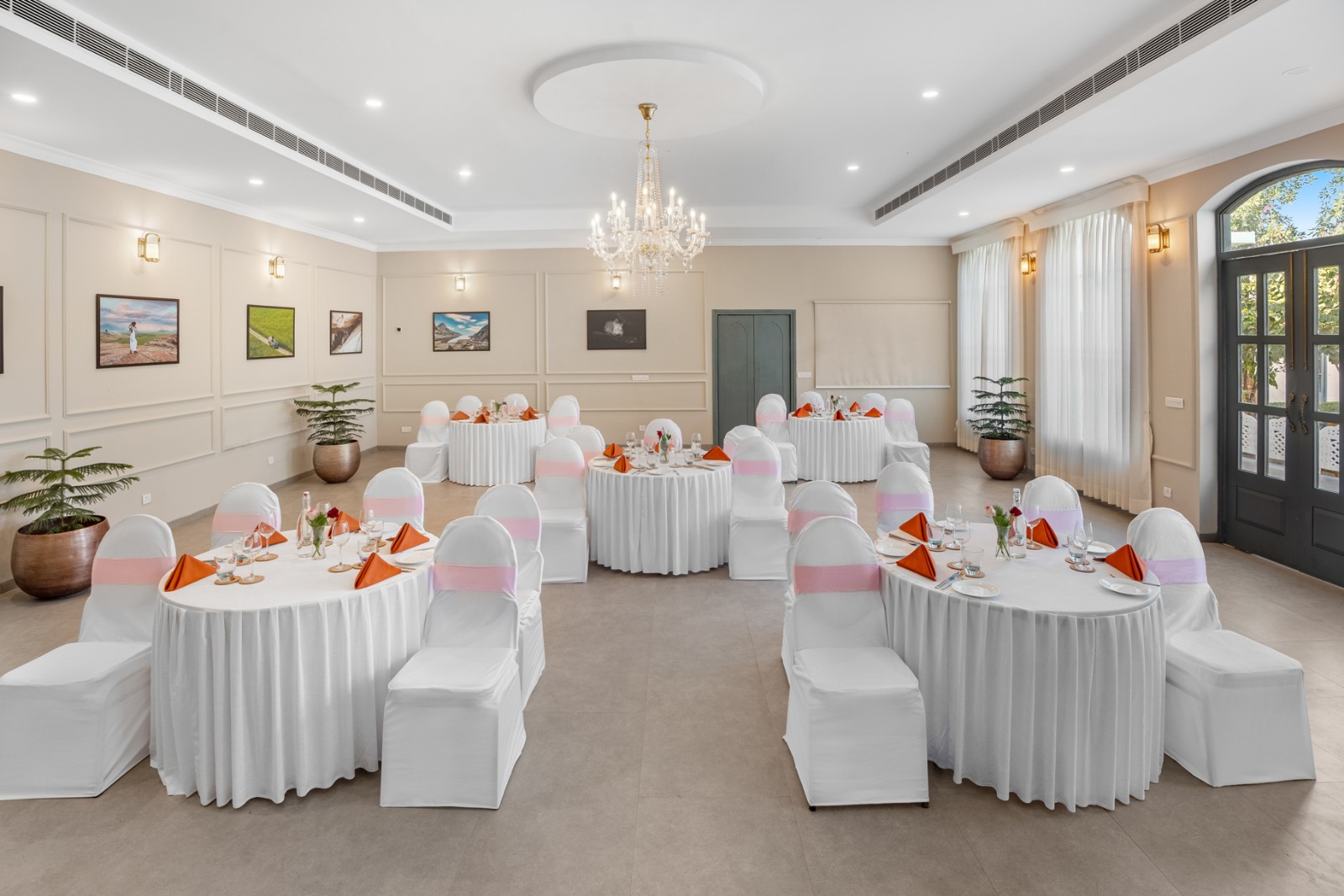 Elegant banquet room set for a celebration with round tables, white chair covers, pink sashes, and orange napkins beneath a crystal chandelier.