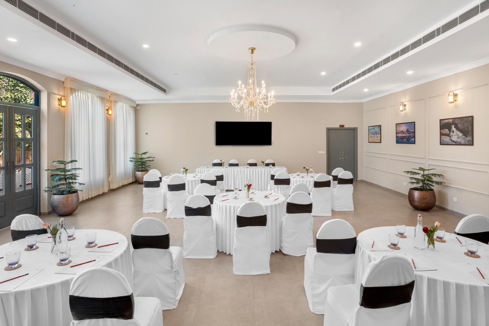Elegant banquet hall set for a conference with round tables, white chair covers, a chandelier, and a wall-mounted screen