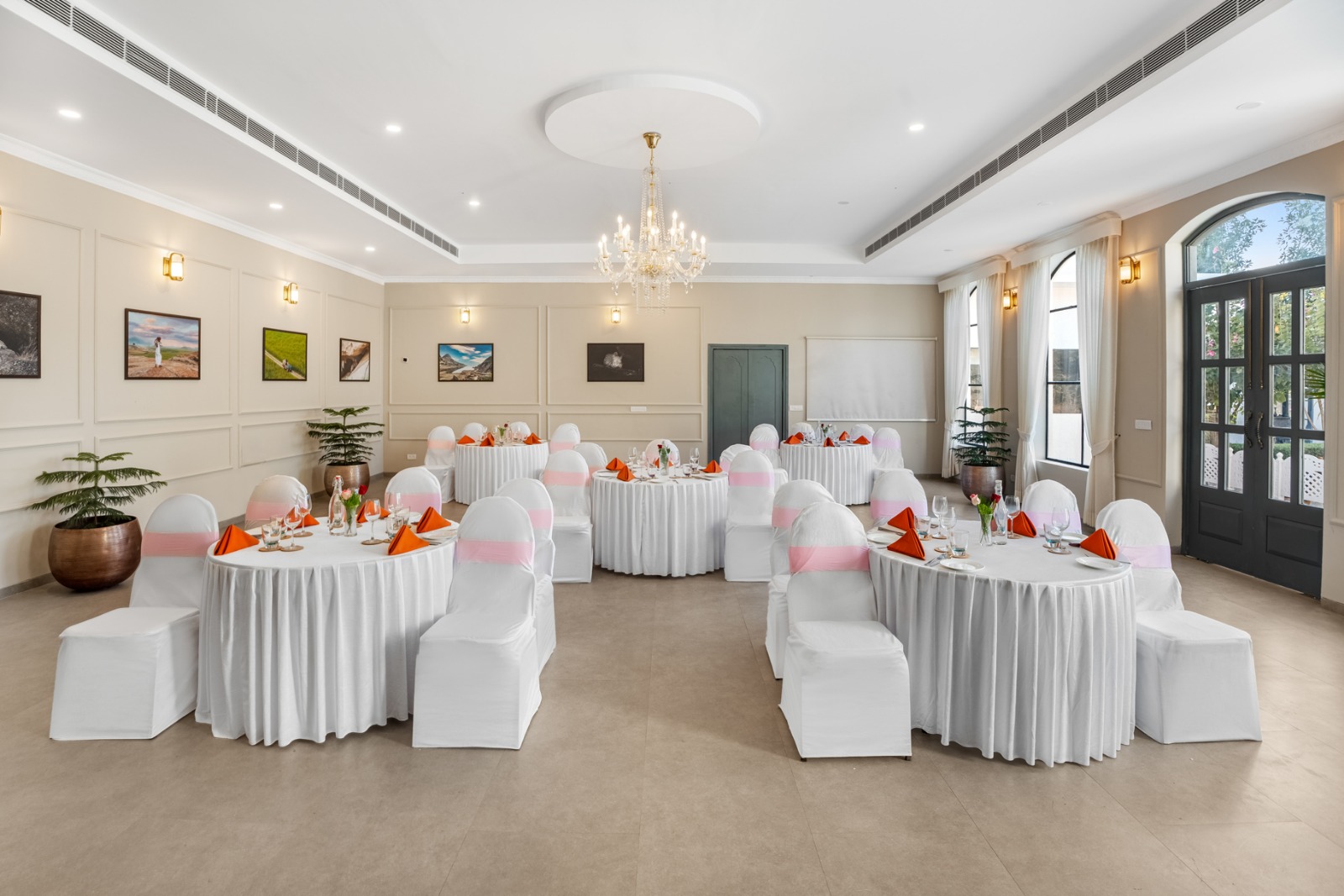 Elegant banquet hall set for a party with round tables, white chair covers with pink sashes, and orange napkins under a crystal chandelier.