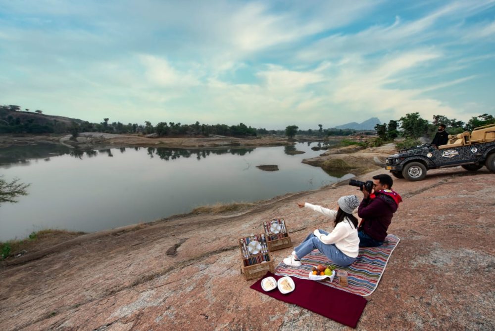 Couple enjoying a riverside picnic on a striped mat with baskets, plates, and fruit, while a photographer shoots the scene; a jeep sits nearby.