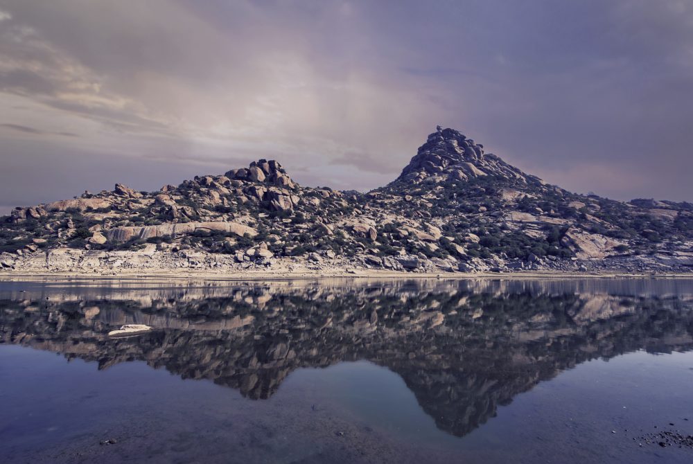 Rugged rocky shoreline with a prominent hill of boulders, reflected in calm water under a purple sunset sky.