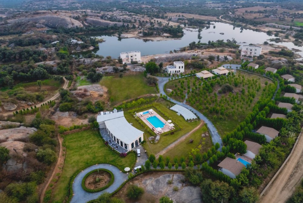 Aerial view of a resort complex with a central curved white building, a blue rectangular swimming pool, and circular driveway surrounded by green lawns and trees near a lake.