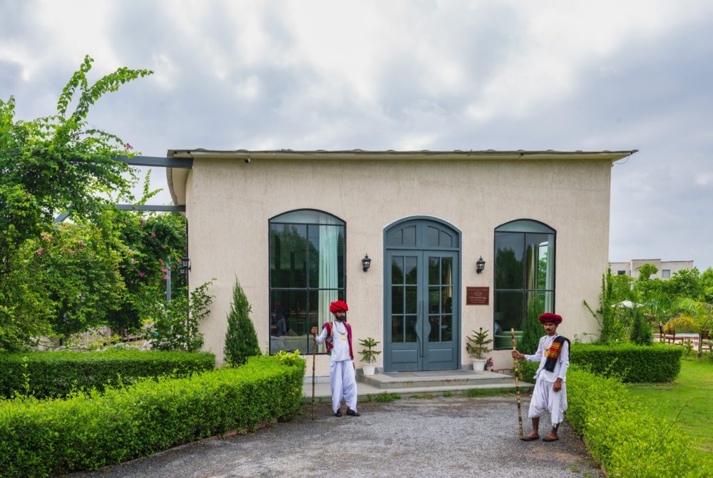 Two men in white robes and red turbans stand with staffs outside a beige building with blue arched doors, surrounded by greenery.