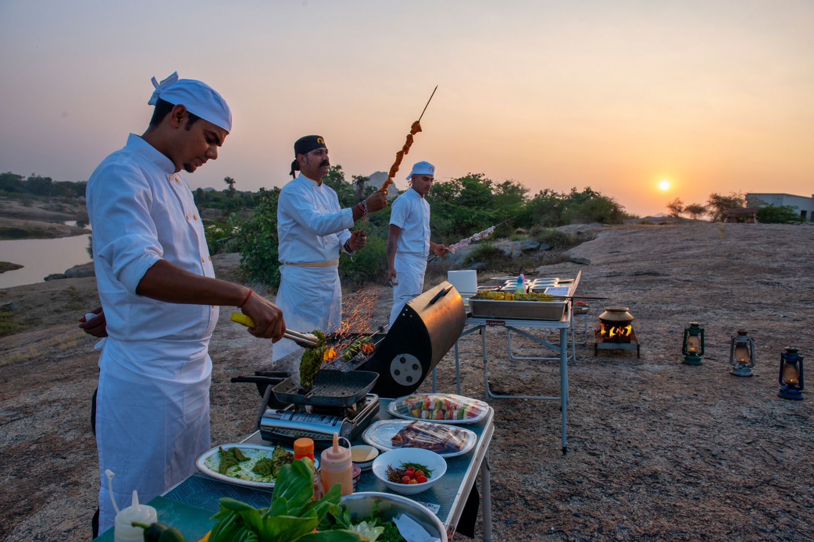 Three chefs in white uniforms grill skewers on a rocky riverside at sunset, with tables of food, sauces, and a portable grill.